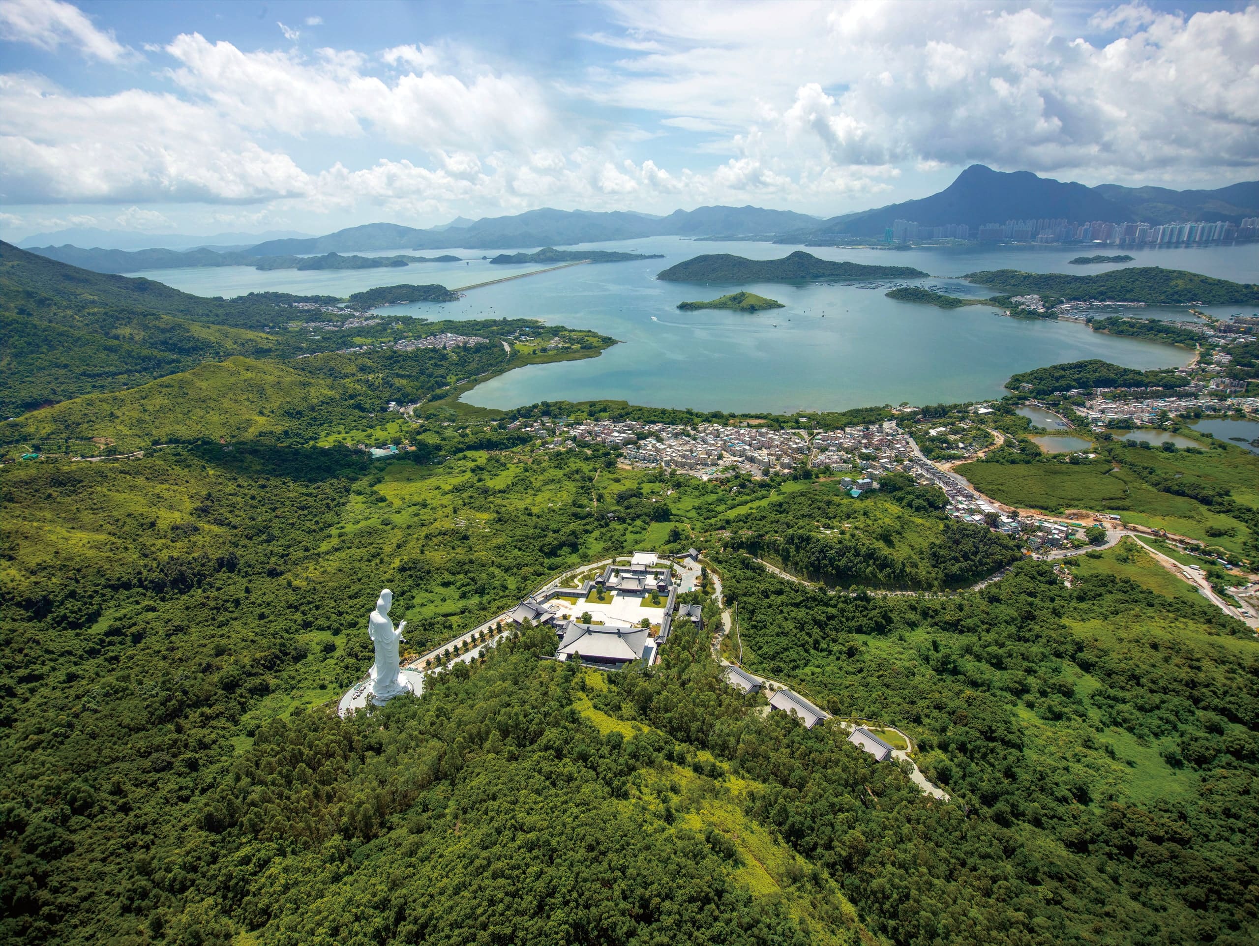Aerial view of Tsz Shan Monastery on a green headland: the white statue at left, tiled halls and a pale courtyard to its right, a road curving up from the village, and the wide water of Tolo Harbour and Plover Cove beyond.