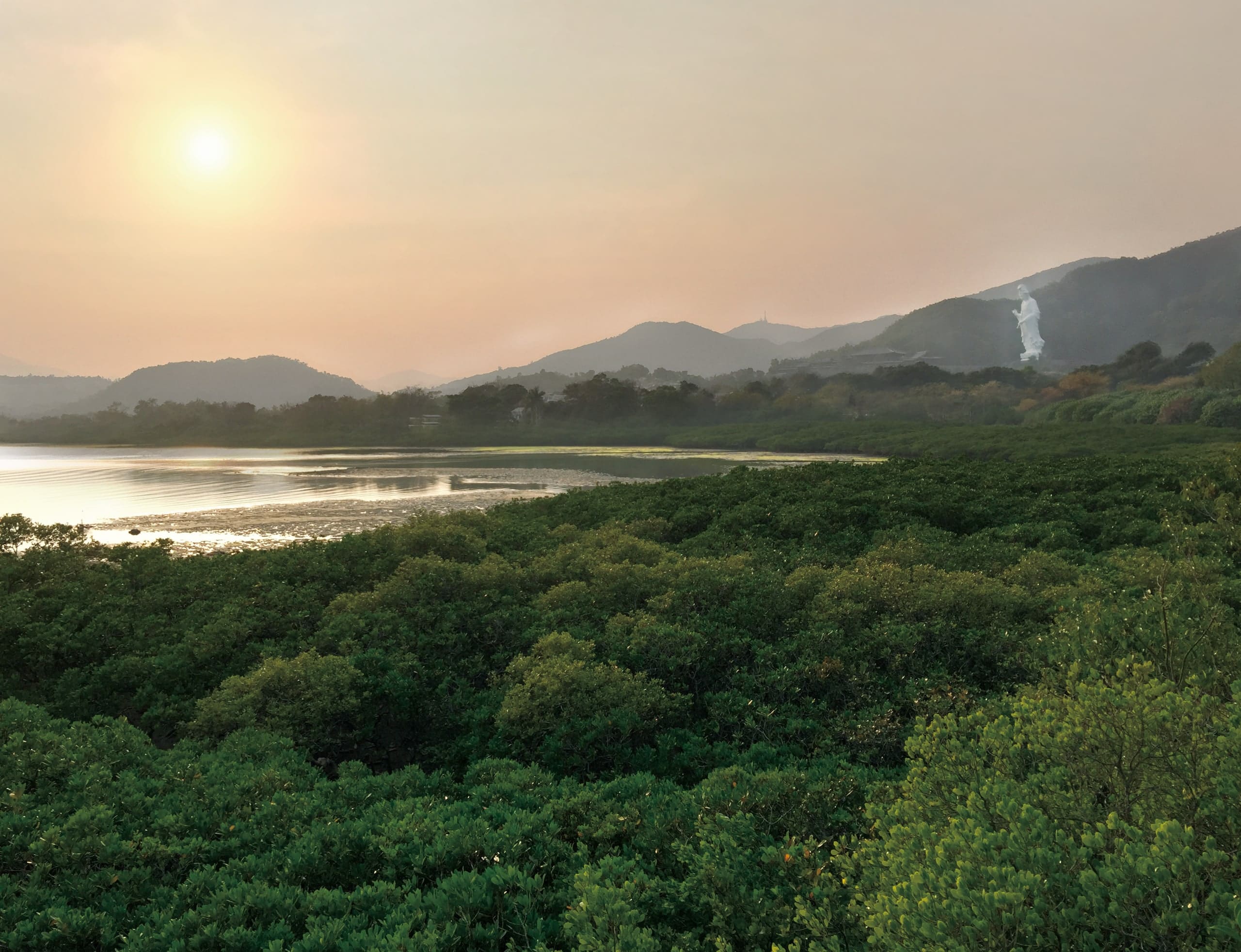 Low sun over a mangrove shore and still water, with a ridge of hills behind and the white statue just visible on the skyline.