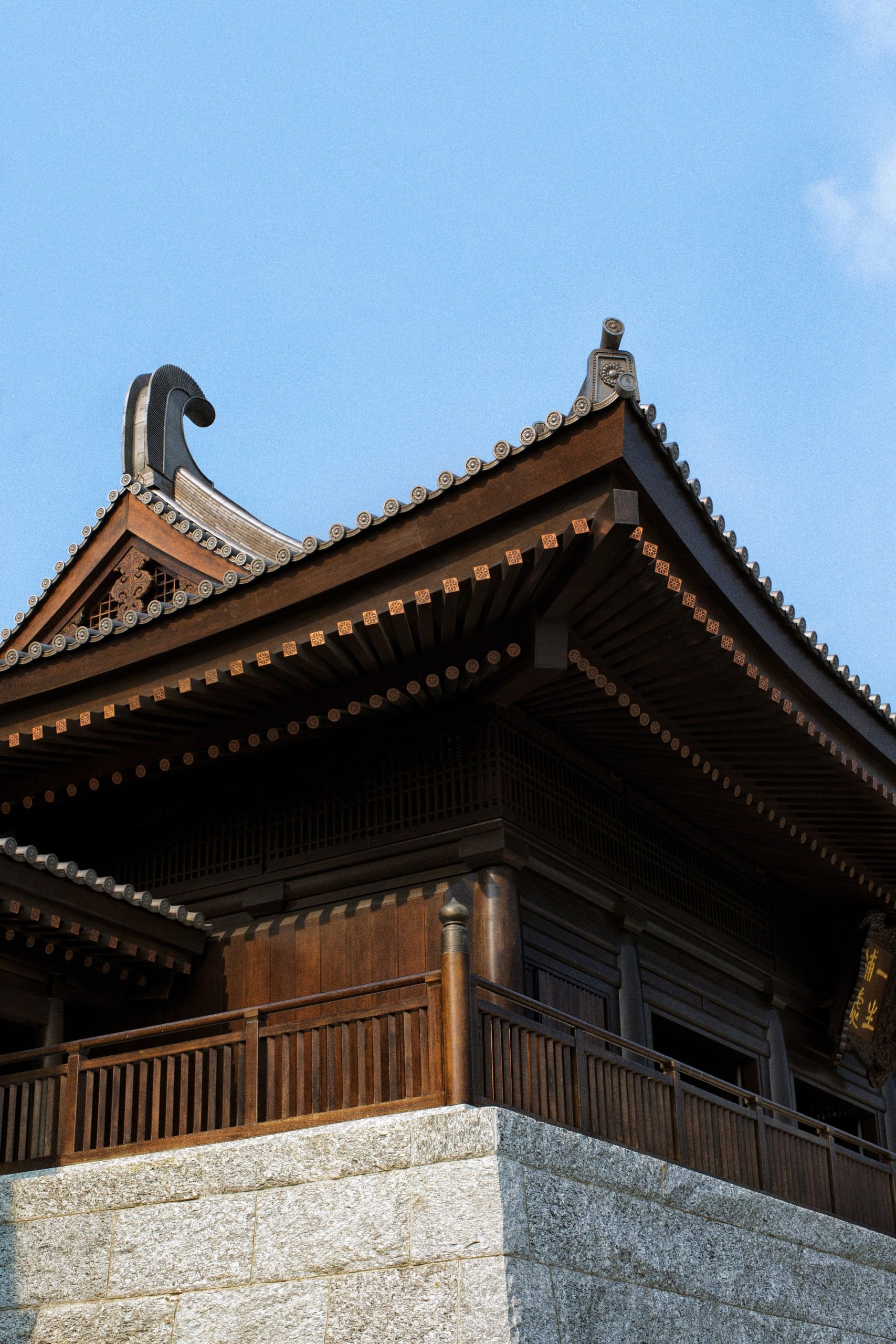 Corner of a timber hall against a cloudless sky: dark bracketed eaves and an upturned ridge ornament above an empty balcony rail, and a pale granite plinth below.