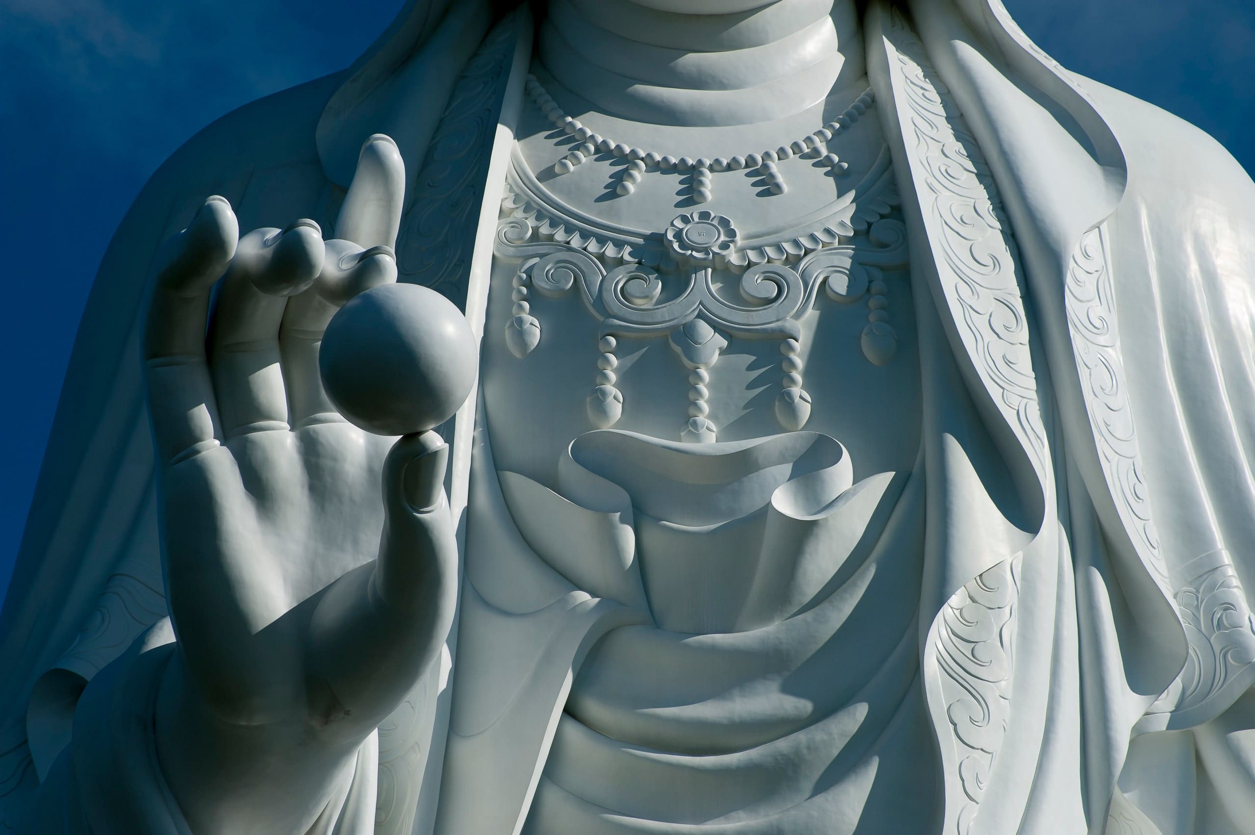 Close view of the Guan Yin statue's raised hand holding a smooth pearl, with carved robe folds and a jewelled pendant behind, against deep blue sky.