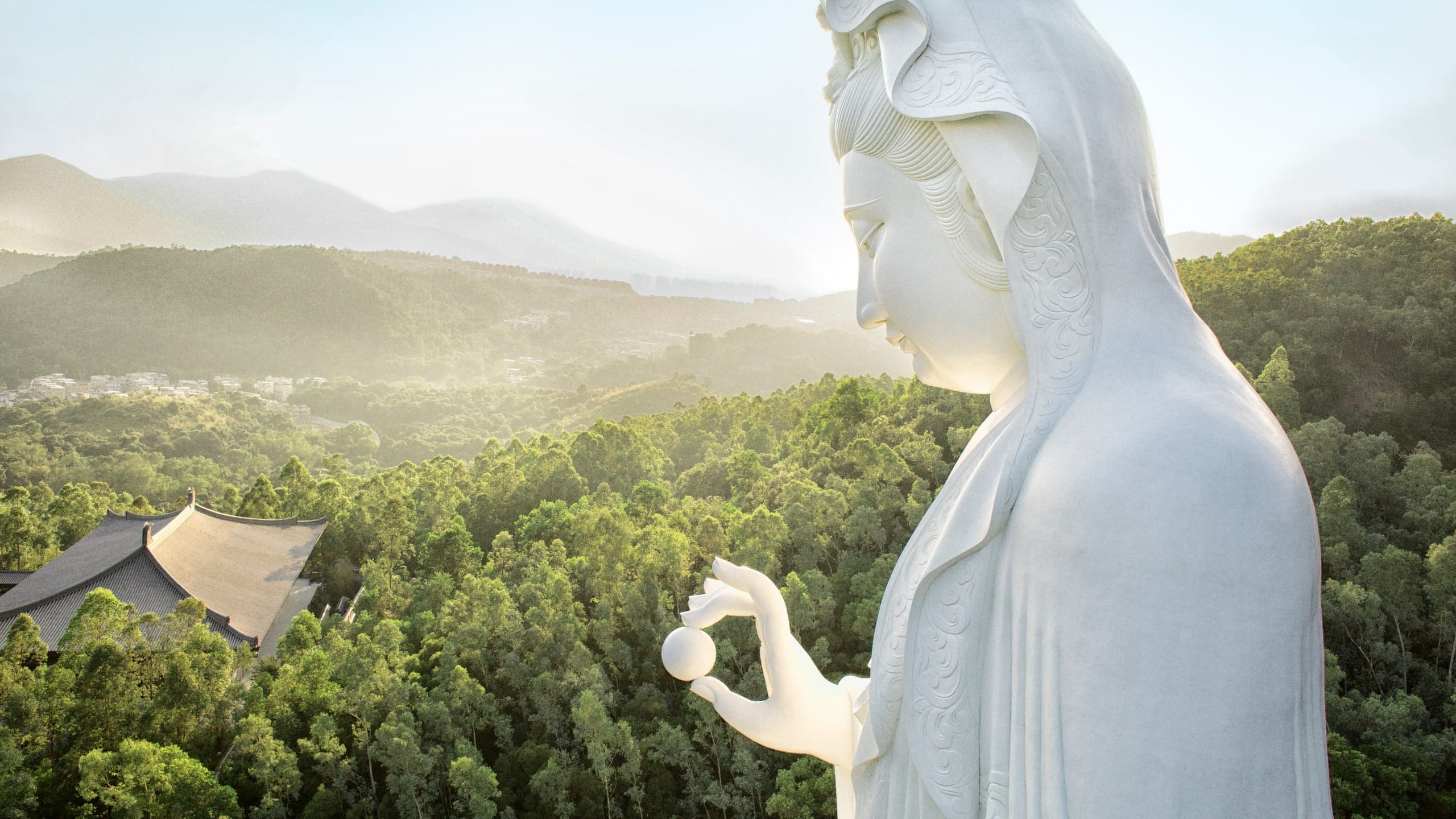 The white Guan Yin statue in profile above wooded hills, holding a small vessel, with mountains fading into morning haze behind.