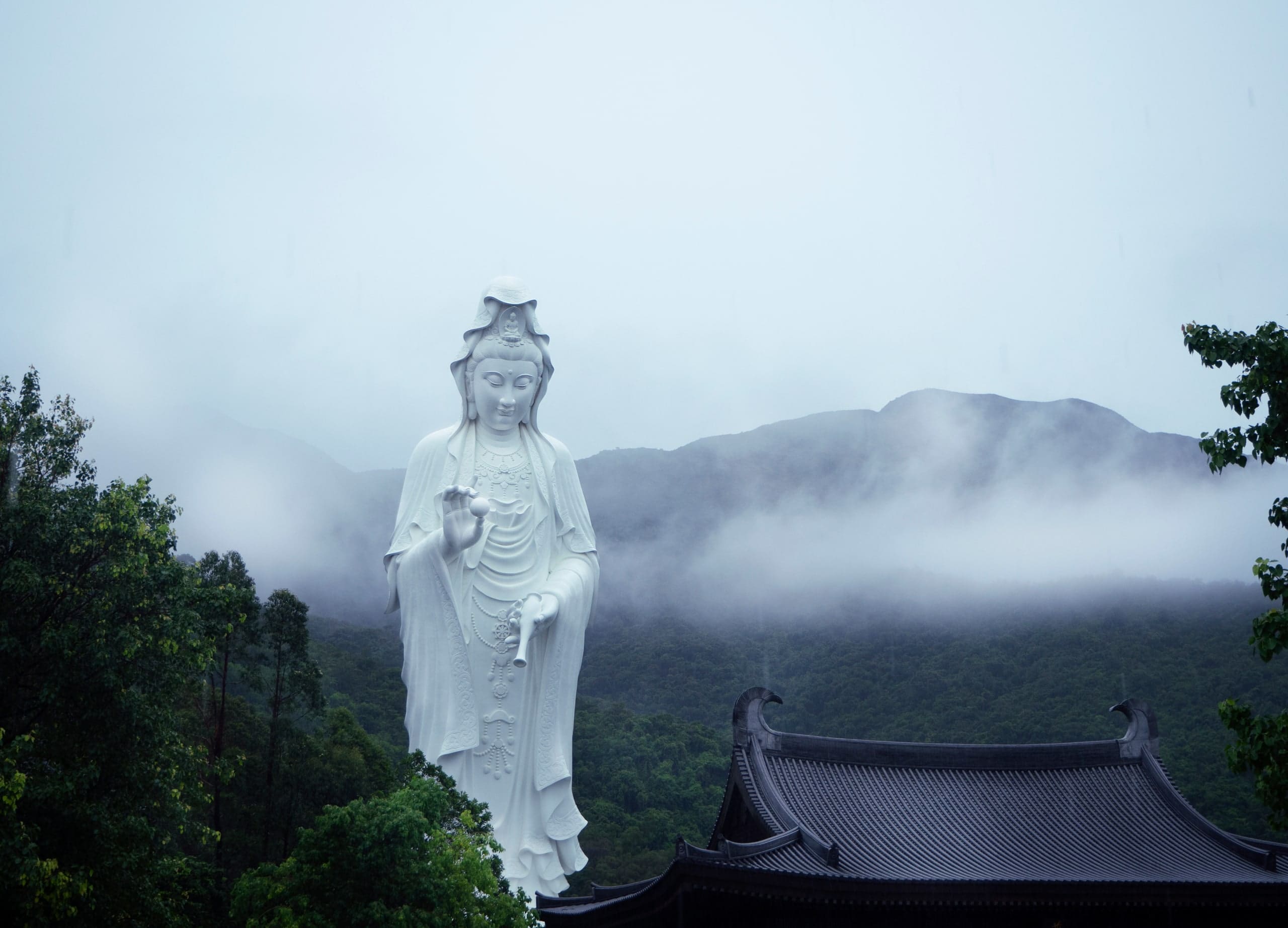The Guan Yin statue standing in low cloud, a dark tiled roof in the foreground and a mountain ridge dissolving into mist behind.