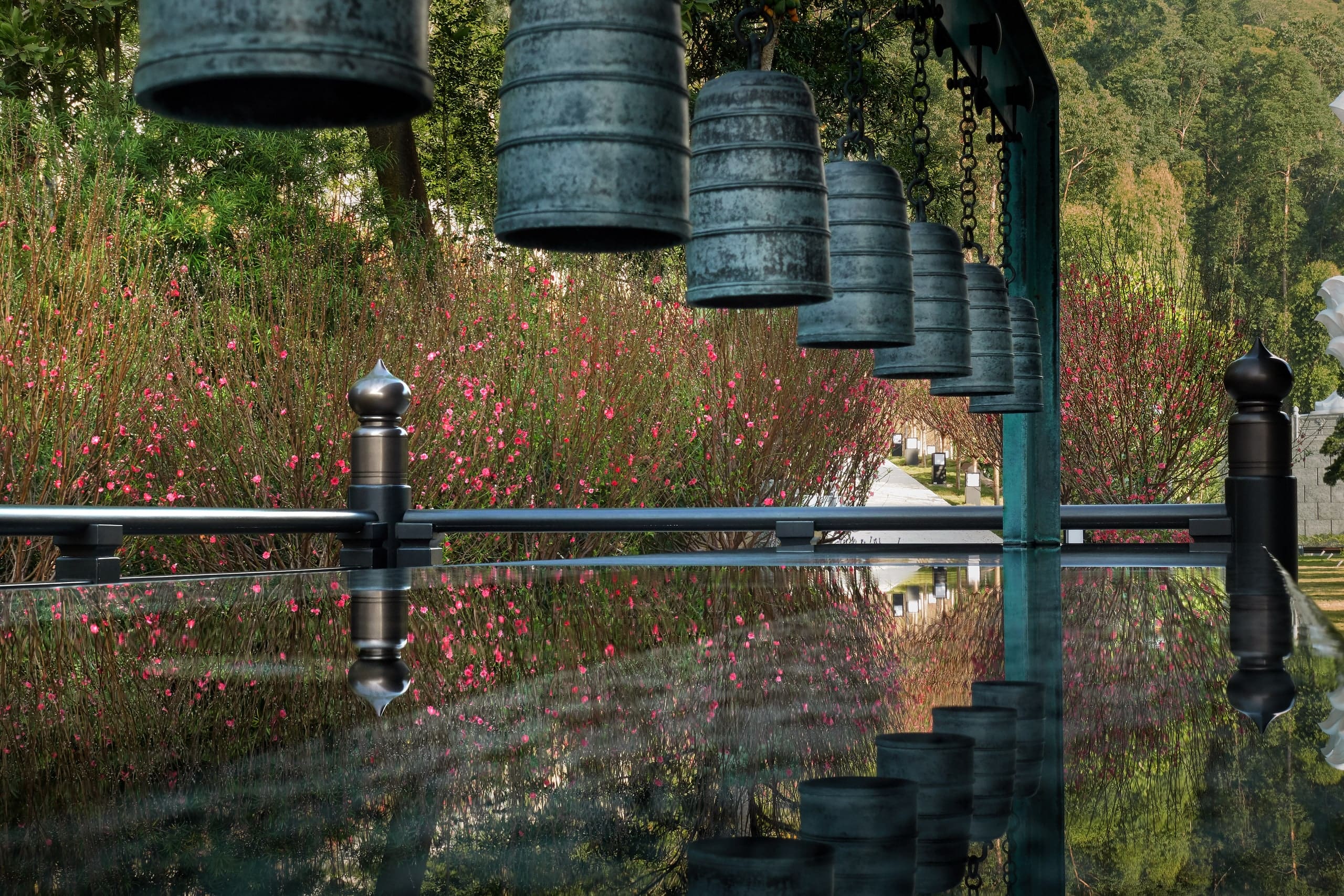 A row of large bronze bells hanging above a still, narrow water channel, with a bank of wildflowers behind and the bells reflected below.